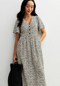 Weißes Kleid mit schwarzen Punkten, kurzen, schwingenden Ärmeln, V-Ausschnitt, Knopfverschluss und gerafftem Taillenbereich. Das Model hält eine schwarze geflochtene Tote-Tasche.