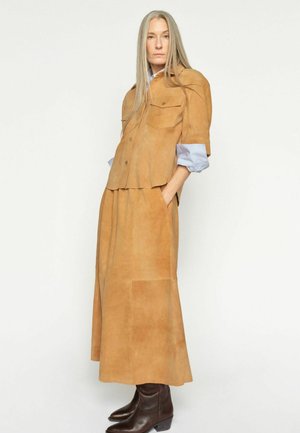 Woman with long gray hair wears tan suede skirt and jacket over light blue shirt, paired with dark brown ankle boots, standing against white wall.