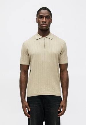 Beige Ripp-Polohemd mit kurzem Kragen und halbzipper, kombiniert mit dunklen Jeans. Das Hemd hat eine strukturierte Stoffoberfläche.