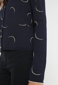 Granatowy kardigan z wzorem srebrnych półksiężyców, posiadający ściągacz przy dole i mankietach oraz małe guziki z przodu.