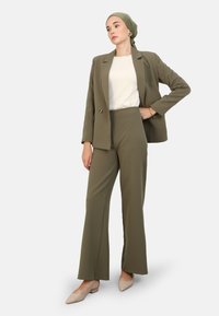 Olive green suit with a tailored blazer and high-waisted flared trousers, paired with a white top and beige shoes. Smooth fabric, minimal accents.