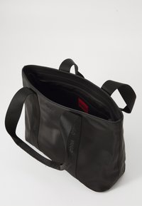 Sac fourre-tout noir en nylon texturé avec des poignées doubles, fermeture éclair et une étiquette rouge à l'intérieur. Présente des détails de logo discrets.