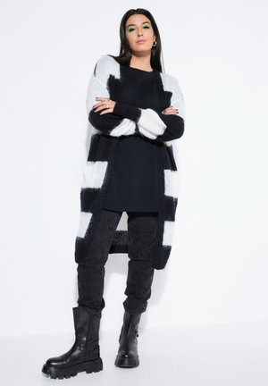 Kvinde stående med armene krydsede iført sort og hvidstribet cardigan, sort kjole, mønstrede bukser og sorte kraftige støvler.