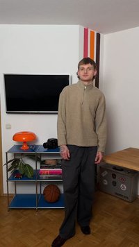 Mężczyzna w beżowej polarze i ciemnych spodniach stoi obok telewizora, pomarańczowej lampy, piłki do koszykówki oraz półki z książkami i aparatem w minimalistycznym rogu pokoju.