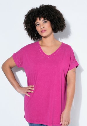 Nainen kiharat hiukset, suuria korviksia ja kirkkaan vaaleanpunainen V-kaula-aukollinen t-paita, käsi lanteilla, seisomassa valkoista taustaa vasten.