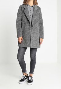 Grå uldblandingsfrakke med notched lapels, enkelt knaplukning, to sidelommer, båret over en grå polo og sorte skinny jeans, parret med sorte sneakers.