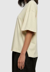 Camiseta de gran tamaño en color beige con mangas cortas, hecha de algodón suave, que presenta una silueta relajada y aberturas laterales para facilitar el movimiento.