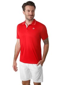 Rotes Poloshirt mit einem weißen Kragen, versehen mit einem kleinen Logo auf der linken Brust. Kombiniert mit weißen Shorts, die ein enges Design zeigen.