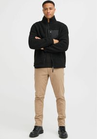 Black fleece jacket with high collar and textured surface, featuring a black zippered pocket. Paired with beige pants and black boots.
