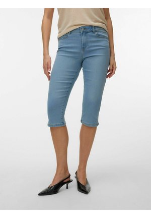 Femeie purtând șorturi din denim albastru deschis, până la genunchi, și pantofi cu toc negri, cu vârf ascuțit și bretea în spate, stând în picioare pe un fond simplu.