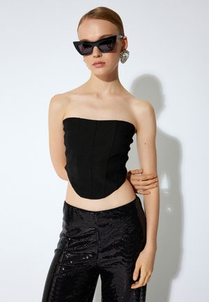 Top negro sin tirantes con paneles texturizados, combinado con pantalones de lentejuelas negros de tiro alto. Modelo llevando gafas de sol negras en forma de ojo de gata y pendientes llamativos.