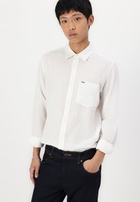 Witte button-up shirt met een gestructureerde afwerking, voorzien van een borstzak en een klein logo. Gecombineerd met donkere spijkerbroek en opgerolde mouwen.