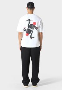 Biała koszulka z graficznym motywem czarnej żurawki oraz czerwonymi okręgami na plecach, zestawiona z czarnymi spodniami i trampkami.