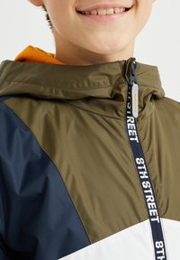 Rompevientos verde y azul marino con un panel blanco, cremallera frontal con una etiqueta de logo y un forro de capucha naranja. Textura ligera y suave.