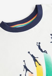 Fehér pamut póló sötétkék, kör alakú nyakkivágással, különböző pózokban elhelyezkedő, árnyékszerű kosárlabdázó játékosokkal. Színes, fokozatosan változó árnyalatú elemek a háttérben.