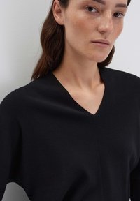 Frau mit leichten Sommersprossen, die einen schwarzen V-Ausschnitt-Pullover trägt und vor einer schlichten hellgrauen Wand posiert.