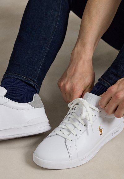 Asmuo riša raiščius ant baltų odinių Polo Ralph Lauren sportbačių, dėvi tamsiai mėlynas džinsines kelnes ir tamsiai mėlynas kojines.