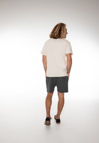 T-shirt beige clair à manches courtes, associé à un short gris foncé, vu de dos. Le sujet se tient sur une surface lisse, portant des chaussures noires à enfiler.