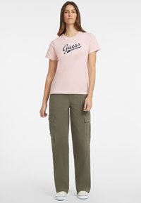 Jasnoróżowa bawełniana koszulka z logo "Guess", połączona z oliwkowymi spodniami bojówkami z dużymi kieszeniami bocznymi i luźnym krojem.