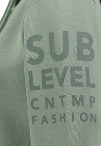 Close-up of muted green fabric with dark green text reading "SUB LEVEL CN TMP FASHION" on a clothing item with a drawstring.