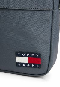 Grå stof rygsæk med en vævet tekstur. Har en rektangulær form og en lap med "TOMMY JEANS" i hvid og rød med marineblå detaljer.