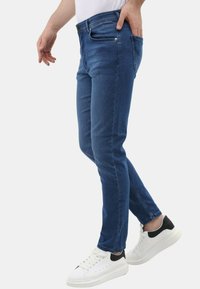 Man in slim-fit blauwe jeans en witte sneakers met zwarte hiel, staand met één hand in de pocket tegen een effen achtergrond.