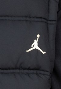 Doudoune marine avec des sections matelassées et un logo Jumpman blanc brodé très visible. Texture lisse avec un léger éclat.
