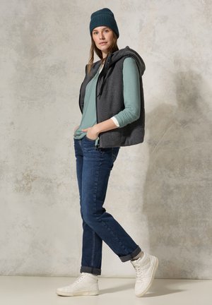 Grijze fleece vest met capuchon over een teal lange mouw shirt, gecombineerd met donkerdenim jeans en witte high-top sneakers. Gebreide beanie.