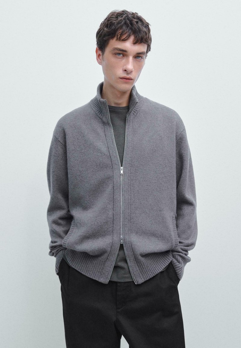 Cardigan grigio in maglia con zip, colletto e polsini a coste, cuciture dettagliate e tasche laterali. Indossato sopra una camicia scura e pantaloni neri.