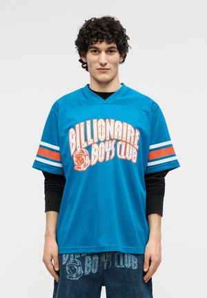 Tânăr cu păr creț, purtând un tricou plasă albastru „Billionaire Boys Club” peste un tricou negru cu mâneci lungi și blugi asortați cu imprimeu.