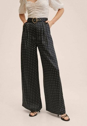 Femme portant un pantalon noir à jambes larges à pois blancs, un haut blanc à manches bouffantes à pois blancs assorti, et des sandales noires à talons.