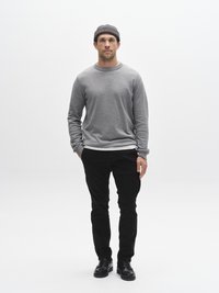 Szürke pamut pulóver kerek nyakkivágással, fekete, szűk szabású nadrág és fekete cipő. A modell szürke kötött sapkát visel. Egyszínű háttér.