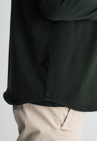 Dunkelgrünes Langarmshirt mit abgerundetem Saum und sichtbaren Nähdetails, mit einem Stoffetikett an der Seite. Kombiniert mit hellbeigen Hosen.