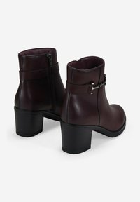 Botas de tobillo de cuero marrón oscuro con cremalleras laterales, correa decorativa con detalle metálico y tacones de bloque sobre un fondo blanco.