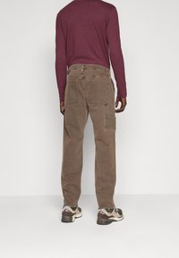 Bruna cargobyxor med flera fickor, en avslappnad passform och ett mjukt, texturerat tyg. Bärs tillsammans med en långärmad vinröd skjorta och sneakers.