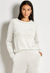 Frau mit langen dunklen Haaren, die goldene Ohringe trägt, ein weißes strukturiertes Langarmoberteil und hellfarbige Hosen, mit den Händen in den Taschen.