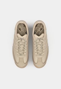 Bež tenisice s glatkom kožnom gornjom strukturom, okruglim vrhom, šest rupa za vezanje, teksturiranim detaljima i "adidas" brendom na jeziku i bočnim stranama.