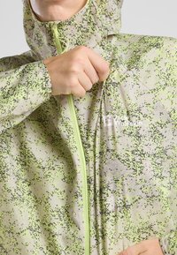 Veste légère et translucide de couleur verte avec un motif à pois noirs. Elle possède une fermeture éclair à l'avant et une capuche réglable pour un ajustement sécurisé.