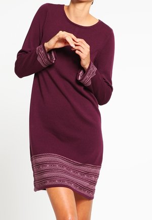 Femme portant une robe en maille violette à manches longues avec des poignets et un ourlet à motifs, debout devant un fond blanc uni.