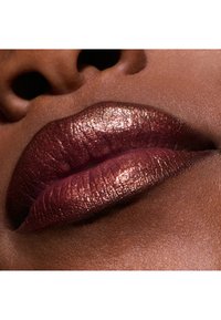 Rouge à lèvres en bronze métallique sur des lèvres bien pulpeuses, avec une base bourgogne profonde et un subtil éclat. Texture lisse sur fond de ton de peau chaud.