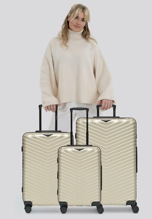 Femme en pull beige tenant les poignées de trois valises rigides dorées avec des roues noires et des poignées télescopiques.
