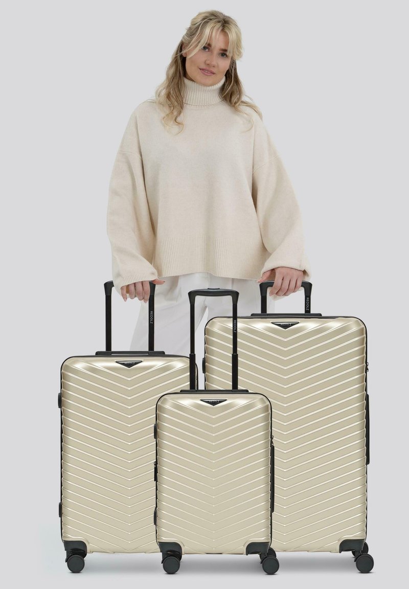 Donna in maglione beige che tiene le maniglie di tre valigie rigide dorate con ruote nere e manici telescopici.