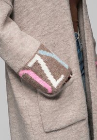 Cardigan in lana beige con vestibilità ampia. I polsini presentano motivi geometrici in marrone, rosa e blu. La maglia strutturata aggiunge calore.