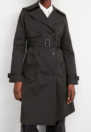 Personne portant un trench-coat noir croisé avec ceinture, poignets à boutons, épaulettes et poches latérales inclinées, par-dessus une chemise blanche à col.