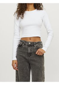 Frau mit lockigem Haar, die ein weißes, langärmliges Crop-Top und hochgeschnittene, ausgebleichte schwarze Jeans trägt, mit der Hand in der Nähe der Tasche.