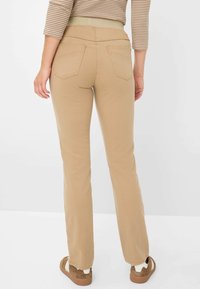 Beige broek met een gladde textuur, voorzien van een brede elastische tailleband en twee achterzakken. De pijpen zijn recht en licht uitlopend.