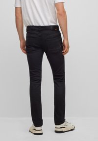 Hombre con jeans ajustados negros, zapatillas blancas y una camiseta blanca, de pie contra un fondo gris claro liso.