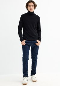 Suéter negro de cuello alto con mangas largas, combinado con jeans azul oscuro y zapatillas blancas con acentos negros y suelas de goma.