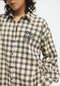 Checkered beige shirt made of a soft fabric, featuring black buttons and an embroidered design on the chest with a mix of brown and blue lines.