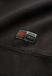 Tela negra con una etiqueta visible. La etiqueta presenta la marca "Superdry" en blanco sobre un fondo verde, con acentos circulares rojos.